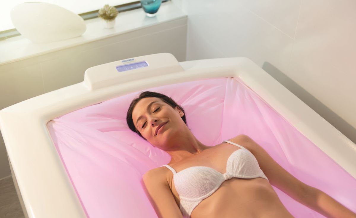 Jeune femme détendue, les yeux fermés, allongée confortablement sur une table d'hydrothérapie ou un caisson de flottaison aux tons rosés dans un centre de bien-être Valdys