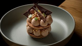 Dessert en couches sophistiqué : deux disques de macaron croustillant garnis de généreuses noisettes de mousse au chocolat crémeuse, surmontés de copeaux de chocolat noir, de noisettes entières grillées et de micropousses, le tout présenté dans une assiet