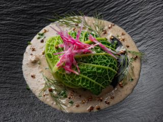 Plat gastronomique végétarien : une ballotine de chou vert frisé farci trônant sur une onctueuse sauce crémeuse, décorée de fins bâtonnets de radis roses et d'herbes fraîches, sur une assiette ardoise