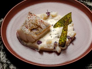 Plat gastronomique composé d'un filet de poisson à la peau croustillante, épicé et grillé, posé sur une purée de chou-fleur onctueuse. L'assiette est décorée de sommités de chou-fleur cru, de deux chips d'algue nori croustillantes et de micropousses pourp